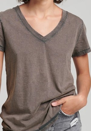 Personne portant un T-shirt marron délavé en col V avec une main dans la poche d’un short en jean gris usé.