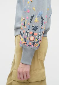 Gaiši pelēks džemperis ar krāsainiem ziedu izšuvumiem uz piedurknes. Ribota aproce ar mīkstu tekstūru; apvienots ar brūniem biksēm.