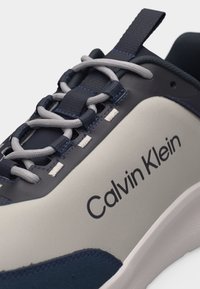 Κοντινή φωτογραφία ενός γκρι και ναυτικού αθλητικού παπουτσιού με γκρι κορδόνια και λογότυπο "Calvin Klein" στην πλευρά, που αναδεικνύει τις θηλιές των κορδονιών και την θηλιά στην φτέρνα.