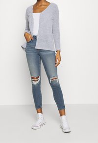 Osoba ubrana w jasnoszary, fakturowany kardigan na białej bluzce, w podarte niebieskie wąskie dżinsy i białe sneakersy za kostkę, stoi z jedną ręką w kieszeni.