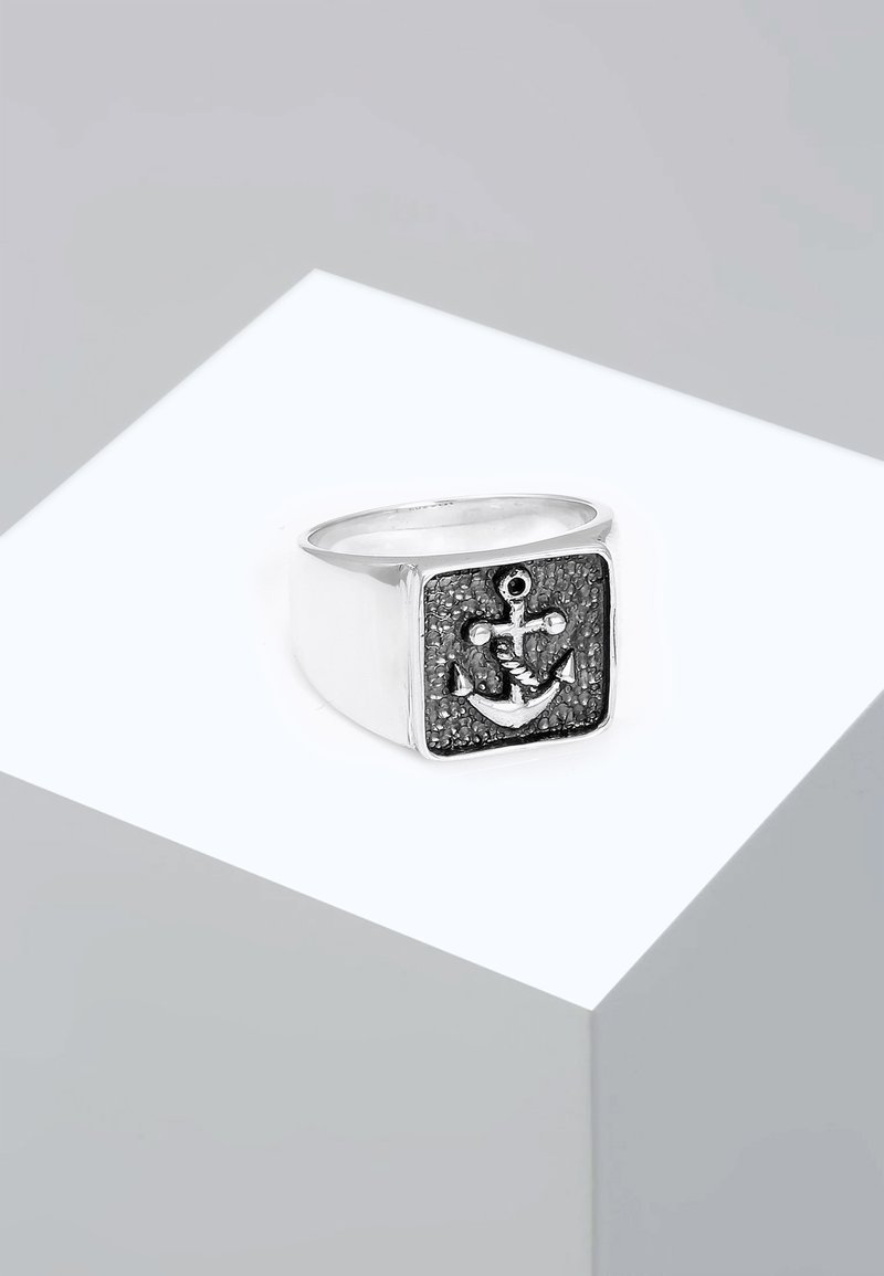Sølv firkantet ring med et tekstureret sort ankerdesign og en lille rund accent, sat mod en glat poleret ring.