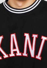 Close-up of black sweatshirt collar with white and black stripes above large white and red embroidered letters "KANI".