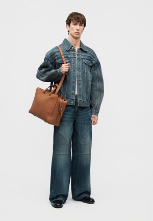 Jeune homme portant une veste en jean surdimensionnée et un jean à jambes larges, tenant un grand sac fourre-tout en cuir marron avec une bandoulière.