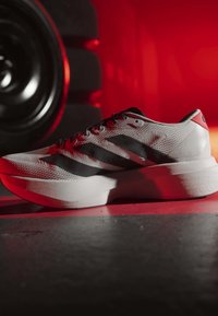 Weißer und schwarzer Sportschuh mit gepolsterter Sohle neben einem großen schwarzen Reifen, beleuchtet von rotem und orangefarbenem Licht in einem Fitnessstudio.