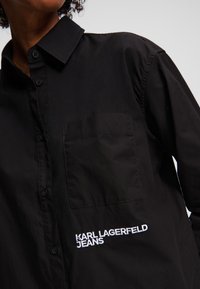 Juodas marškinis su mygtukais, su smailiu apykaklės dizainu, priekyje yra kišenė, o kairėje pusėje siuvinėtas baltas logotipas "KARL LAGERFELD JEANS".