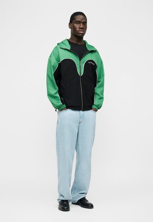 Mladý muž stojaci s rukami v kapsách svetlomodrých riflí, oblečený v čierno-zelené bundě na zips a čiernych topánkach, na bielom pozadí.