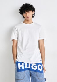 Camiseta de algodón blanca con cuello redondo, combinada con unos pantalones cortos de denim azul que tienen un gran texto blanco "HUGO" en la parte trasera.