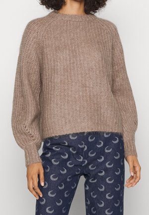 Personne portant un pull ample texturé marron clair et un pantalon bleu foncé avec un motif répétitif de croissant de lune.