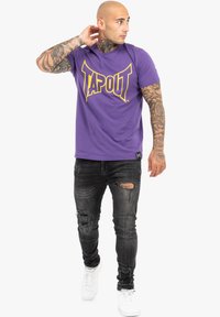 Lilas pamut póló sárga "TAPOUT" logóval, fekete, szakadt farmernadrággal és fehér sportcipőkkel párosítva. Egyszerű háttér.