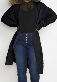 Czarny dziany kardigan o teksturowanym wzorze, noszony na czarnej koronkowej bluzce i niebieskich dżinsach z wysokim stanem, posiadających cztery guziki.