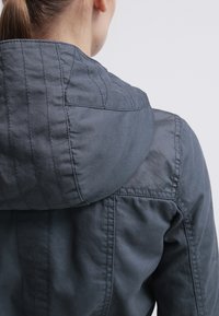 Chaqueta gris con capucha y acabado texturizado, con costuras visibles y un diseño casual, vista desde atrás.