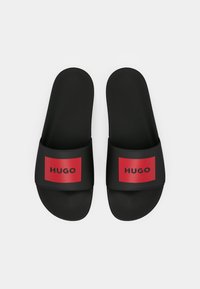 Melni slaidu sandalīši ar platām siksnām, kuru izskatā ir sarkani taisnstūrveida pleķi un melns "HUGO" teksts uz katras siksnas, izstādīti uz baltā fona.