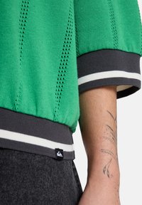 Pull vert en maille avec un design à mailles ouvertes, poignets rayés gris et étiquette logo noire à l'ourlet. Tatouage visible sur la peau du bras.