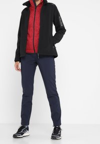 Veste softshell noire sur une polaire rouge zippée, associée à un pantalon bleu marine et des chaussures de sport noires. Les caractéristiques incluent des zips, des poches et un tissu texturé.