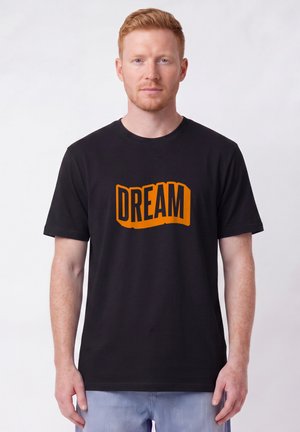 Young man with short red hair wearing black t-shirt with orange text "DREAM" and light blue jeans on plain white background.
