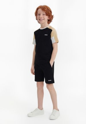 Garçon aux cheveux roux debout, portant un short noir, un t-shirt noir à manches courtes avec des manches beige et blanches, et des baskets blanches.
