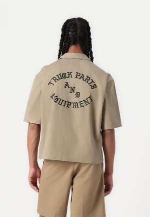 Short-sleeve beige shirt with a relaxed fit, featuring black text on the back that reads “TRUCK PARTS AND EQUIPMENT.” Smooth fabric texture.