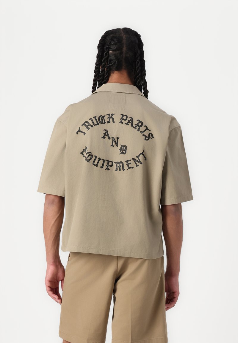 Camicia beige a maniche corte con una vestibilità comoda, con testo nero sul retro che recita "PARTI E ATTREZZATURA PER CAMION". Tessuto dalla trama liscia.