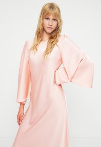 Μακρύ φόρεμα με μανίκια σε σχήμα καμπάνας από απαλό, ανοιχτό ροζ ύφασμα, με στρογγυλή λαιμόκοψη και άνετη γραμμή, λεία υφή και πλαϊνές σχισμές.