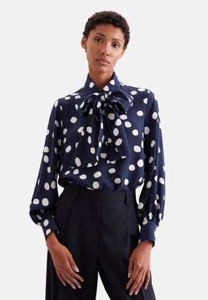 Donna che indossa una camicetta blu navy con grandi pois bianchi e un maxi fiocco al collo, abbinata a pantaloni neri a vita alta.