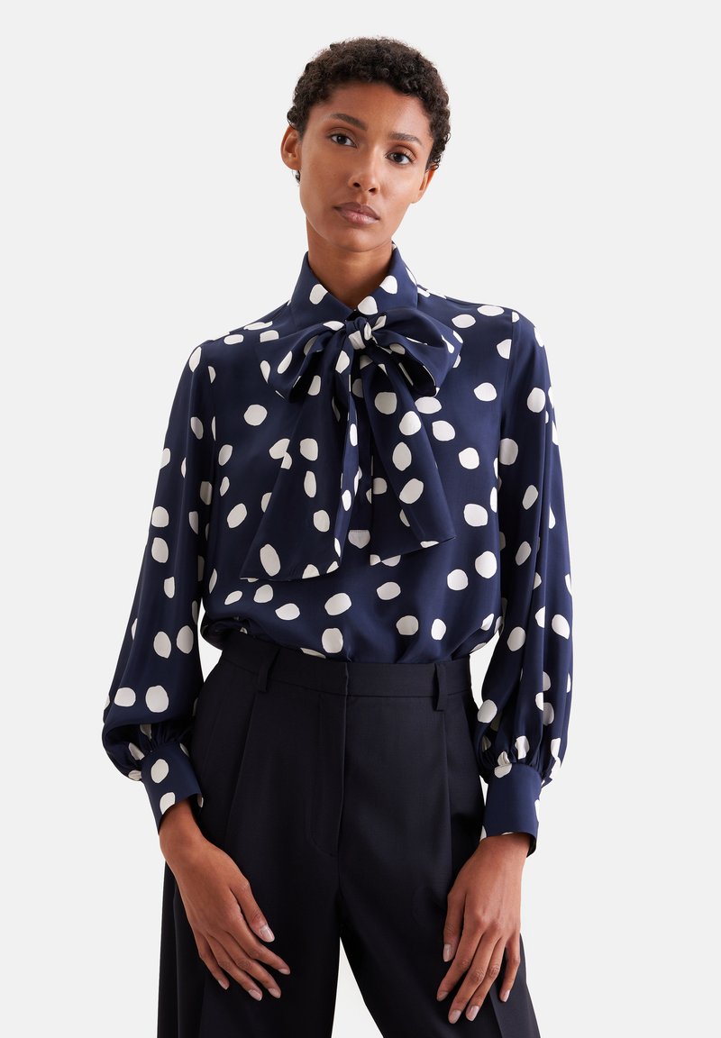 Femme portant un chemisier bleu marine à gros pois blancs et un nœud oversize au col, associé à un pantalon noir taille haute.