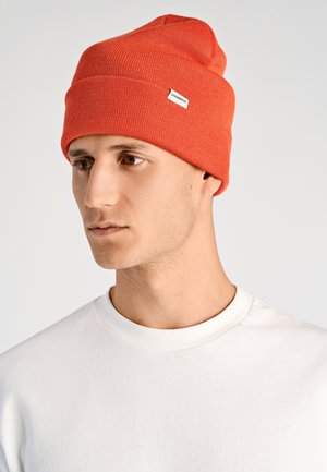 Giovane uomo che indossa una semplice camicia bianca e un brillante berretto lavorato a maglia arancione con una piccola etichetta bianca Lindbergh sul lato.