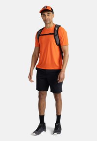 Oranžové sportovní tričko, černé šortky a černé tenisky. Černý batoh s nastavitelnými popruhy a černá s oranžovou čepicí s logem.