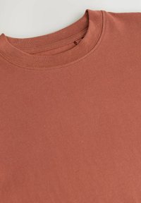 Roestkleurig katoenen T-shirt met een ronde hals. Glad textuur, minimale naden en korte mouwen. Lichtjes gevouwen om de vorm te tonen.