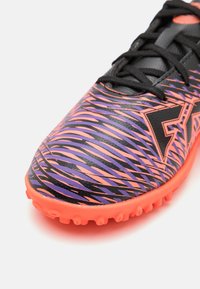 Crampon de football noir et orange avec une tige texturée, présentant un motif en rayures ondulées violettes et roses et une semelle extérieure en caoutchouc moulé.