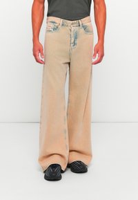 Homme portant un jean beige ample à jambes larges et des baskets noires, debout, les mains détendues le long du corps, sur un fond blanc uni.