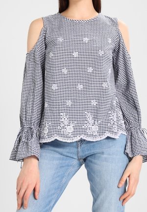 Musta-valkoinen ruudullinen olkapäät paljastava pusero, jossa on kukkakirjailu, hammaslaitainen helma ja levenevät hihansuut, yhdistetty vaaleansiniin farkkuihin.