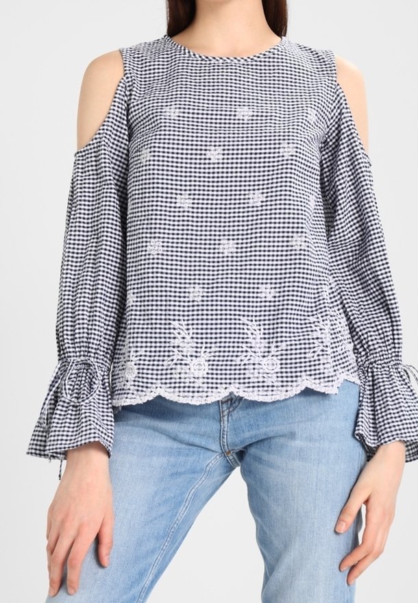 Blouse à épaules dénudées en vichy noir et blanc avec broderies florales, ourlet festonné et poignets évasés, associée à un jean bleu clair.