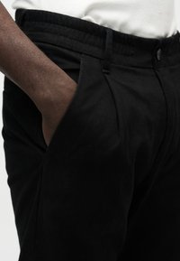 Un pantalon noir fuselé présente un tissu texturé, une fermeture à bouton et des poches latérales, avec une taille élastique pour plus de confort.