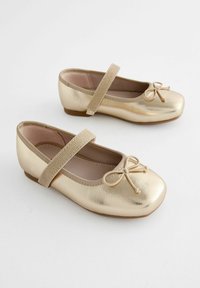 Ballerines métalliques dorées avec un bout arrondi, un détail en nœud et une sangle élastique pour un maintien sécurisé, montées sur une semelle basse marron.