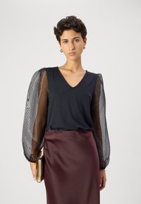 Fekete V-nyakú felső áttetsző, pöttyös puffujjakkal, bordó selyemszoknyával párosítva. A modell egy kis arany clutch-ot tart. Egyszerű, elegáns dizájn.