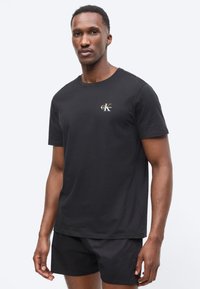 Homme portant un t-shirt noir Calvin Klein et un short noir, debout devant un fond clair uni, face à l'avant avec une expression neutre.