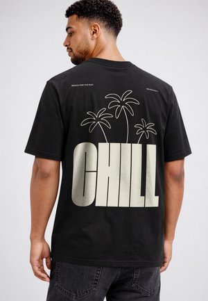 Mann trägt ein schwarzes T-Shirt mit drei Palmen und dem großen Wort "CHILL" auf dem Rücken, steht vor einem einfarbigen Hintergrund.