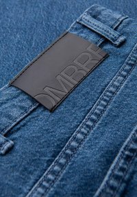 Primer plano de una tela de mezclilla azul con costuras cosidas y una etiqueta de cuero negro embossada con la parte del nombre de la marca "OMBR".