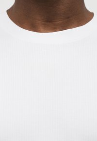 Άτομο με σκουρόχρωμο δέρμα που φοράει λευκό μπλουζάκι με ριμπ ύφανση και στρογγυλή λαιμόκοψη, εμφανίζοντας μόνο το λαιμό και το άνω μέρος του στήθους.