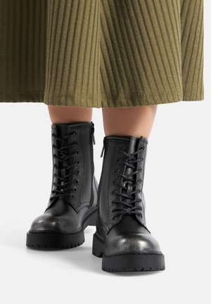 Czarne bojowe botki na sznurowadła noszone z oliwkowo-zieloną prążkowaną spódnicą midi na białym tle.