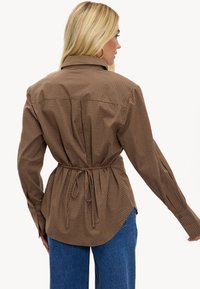 Chemise à carreaux marron avec une taille cintrée, manches longues et coupe décontractée. Dotée d'un col à revers et d'un ourlet arrondi.