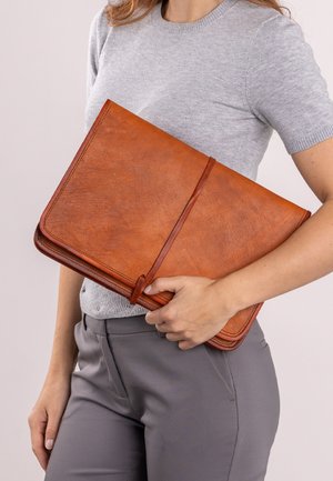 Pochette pour ordinateur portable en cuir marron avec une texture lisse, de forme rectangulaire et une fermeture par sangle en cuir enroulée. Tenue sous le bras.