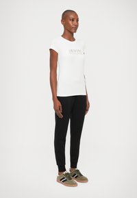 Biely priliehavý tričko s logom "Armani Exchange", v kombinácii s čiernymi teplákmi a olivovými teniskami s čierno-bielymi pruhmi.