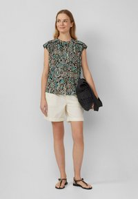 Top senza maniche a fantasia con design geometrici in nero, turchese e beige, abbinato a pantaloni corti crema e con in mano una borsa nera testurizzata.