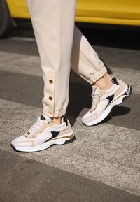 Sneakers bianchi con accenti beige e neri, sovrapposizioni testurizzate e suola spessa. Indossati con joggers beige chiaro con bottoni dorati.