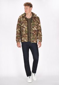 Giacca camouflage in verde, marrone e beige; materiale leggero con chiusura zip e colletto rialzato. Abbinata a una camicia verde e jeans scuri.