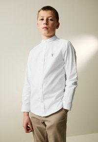 Weißes Hemd mit Stehkragen und einem kleinen blauen Stickdetail, kombiniert mit hellbeigen Hosen.