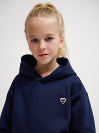 Blondes Mädchen mit blauen Augen, das einen marineblauen Hoodie mit einem kleinen Bienensymbol auf der Brust trägt und vor einem schlichten weißen Hintergrund steht.
