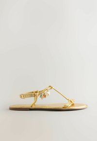 Sandale plate dorée avec bride à la cheville, boucle nouée au niveau des orteils et petits charms suspendus, dont un coquillage et une étoile de mer.