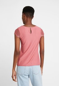 Rosa T-shirt med blonderærmer, detalje med nøglehul i ryggen og blødt stof. Den har en tætsiddende pasform og en glat tekstur.